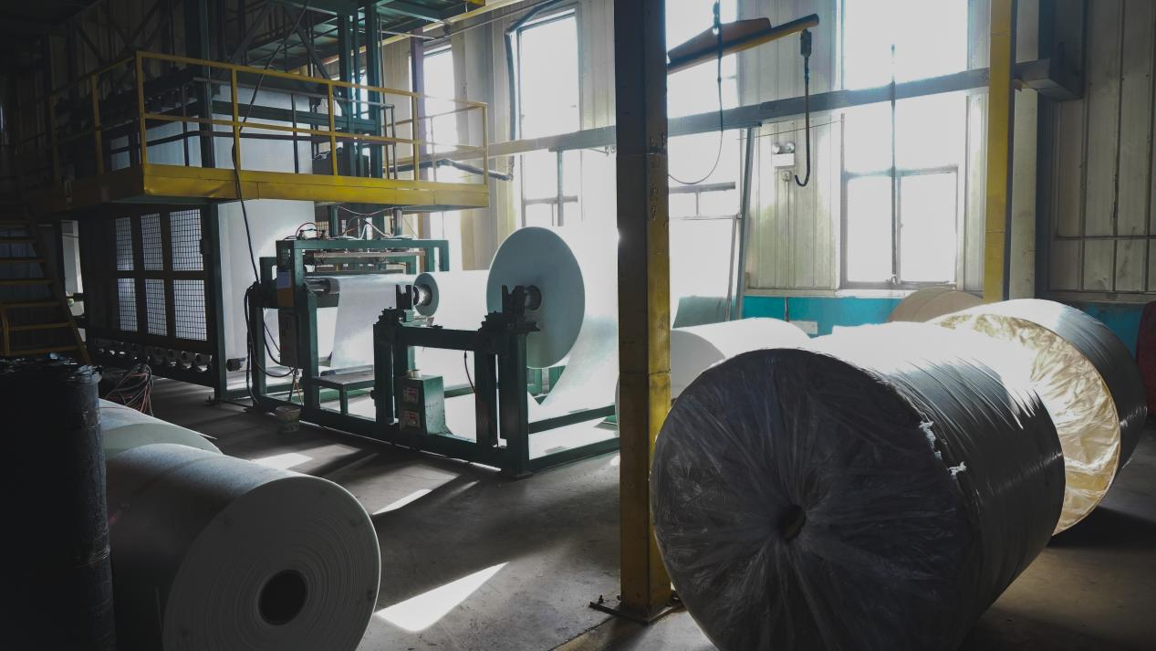 Interior of Attiroof's asphalt shingle manufacturing plant showing rolls of fiberglass mat and the coating machinery used in the production process.