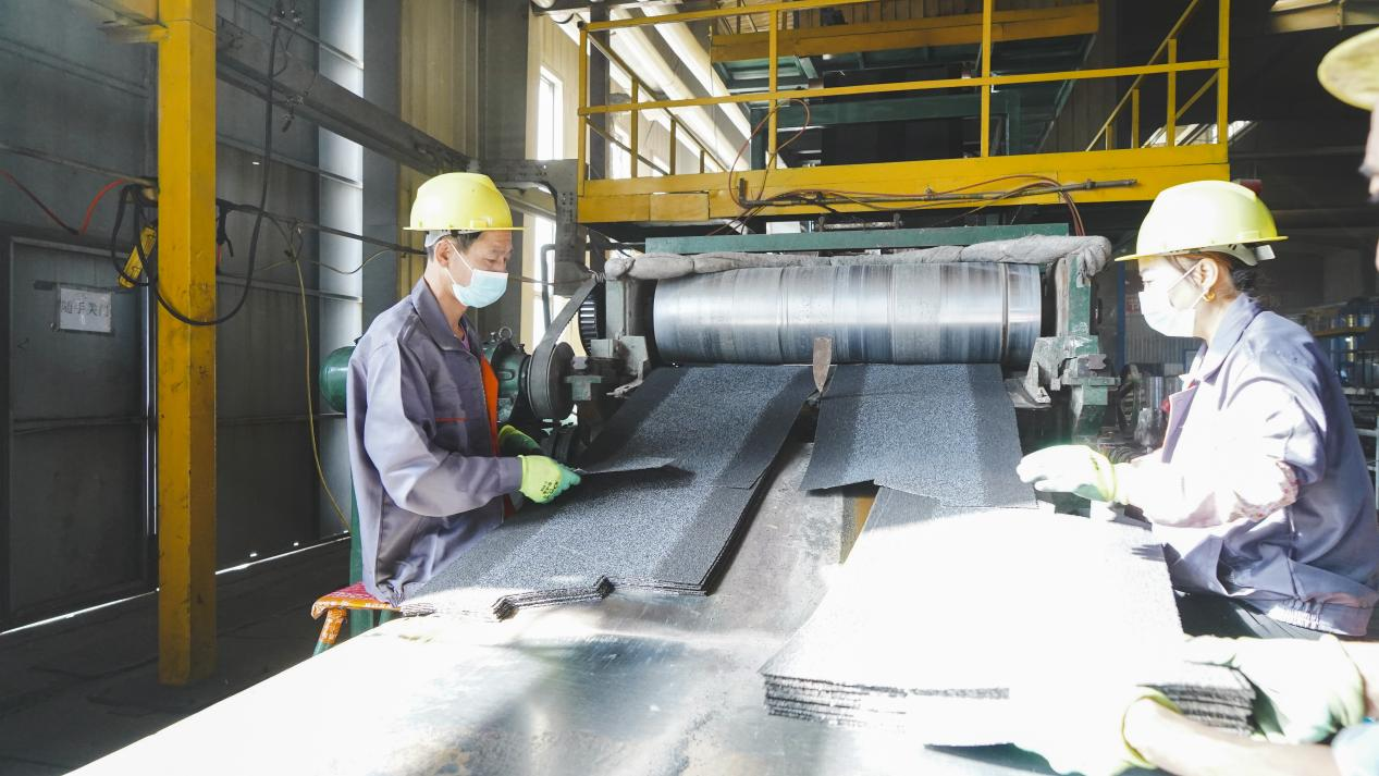 Two workers, dressed in overalls and hard hats, work intently on machinery inside the factory, handling a pile of black asphalt shingles.